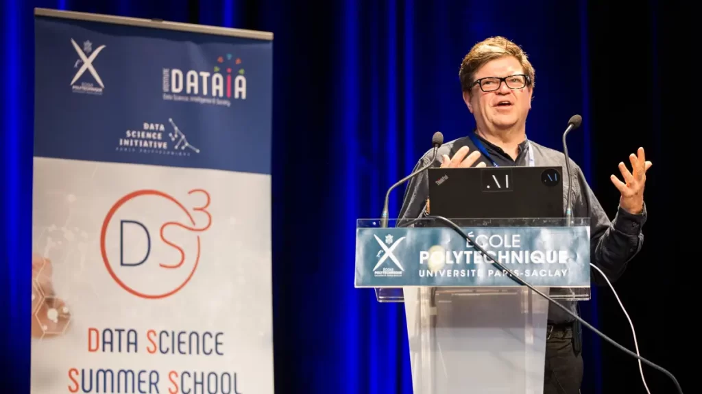 Yann LeCun delivers a keynote at École Polytechnique’s Data Science Summer School, standing at a podium with laptop and microphones on a stage.